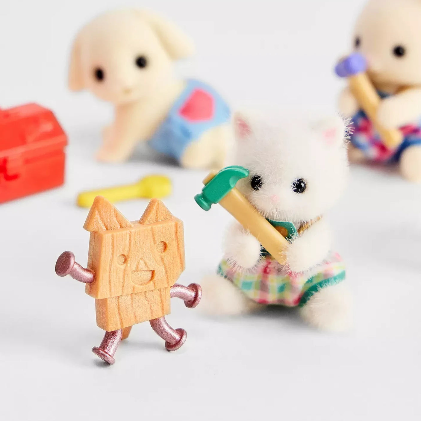 The Sylvanian Families Blind Bag - Baby Builder features a small stuffed cat with a toy hammer beside a wooden figure with nail limbs, with construction toys and a red toolbox, perfect for fans of Sylvanian Families.