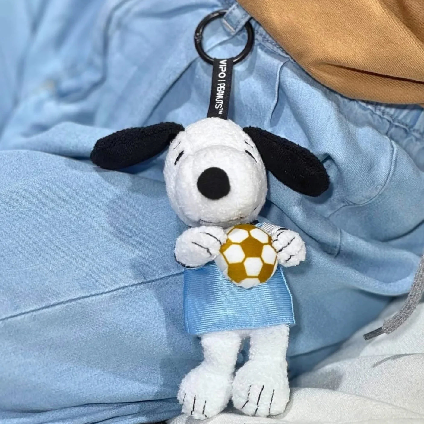 Snoopy soccer plush keychain on light blue fabric, holding a yellow soccer ball