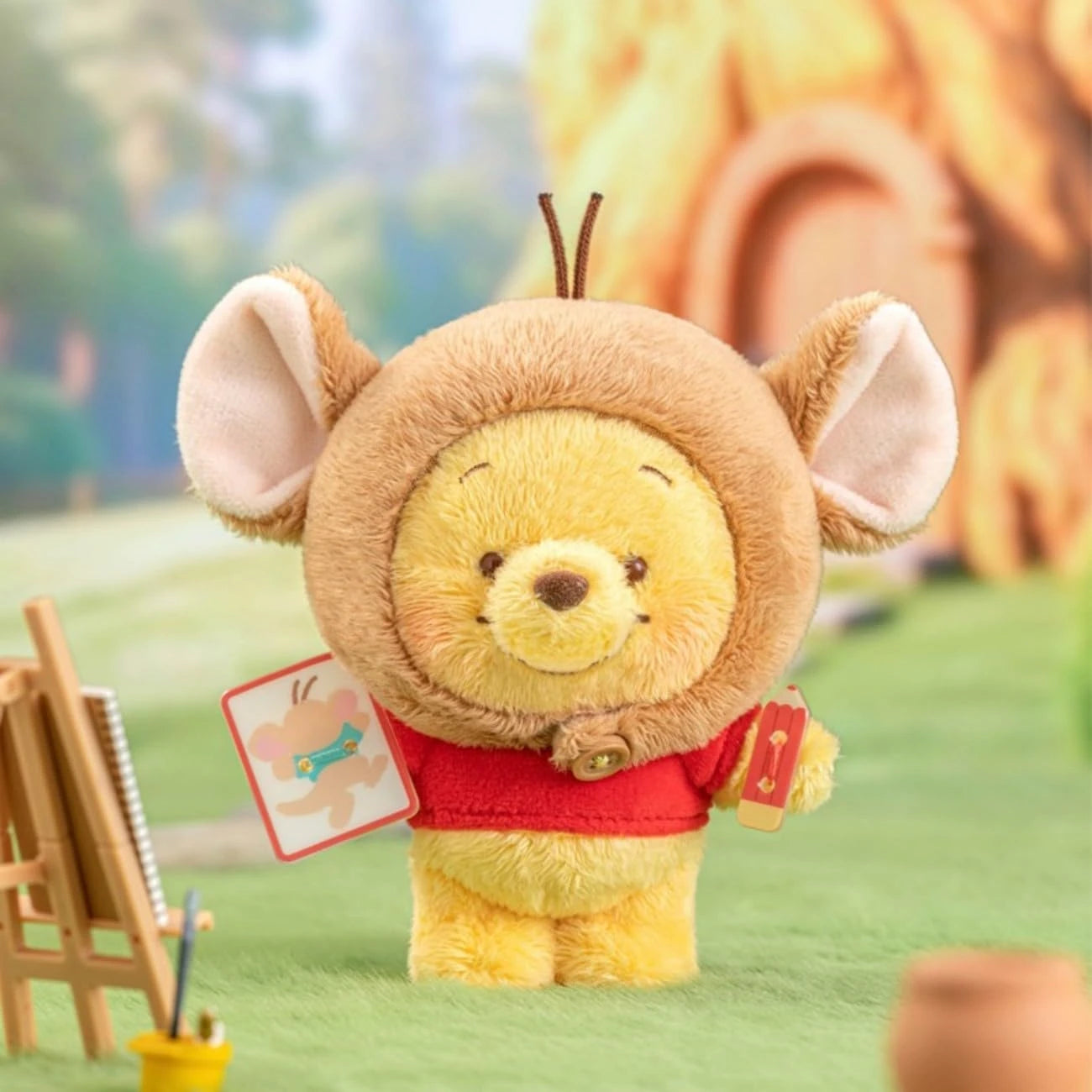 Plush yellow bear wearing a mouse hood and red shirt holding a card and small box outdoors with soft background