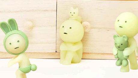 Three pastel-colored Smiski figurines in front of a wooden background. The left figurine is dressed as a bunny, the middle one has a small animal on its head, and the right is holding a teddy bear. These are collectible blind box figures.