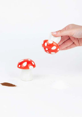 Hand lifting lid off red-and-white spotted mushroom salt shaker with pepper sprinkled nearby on white surface