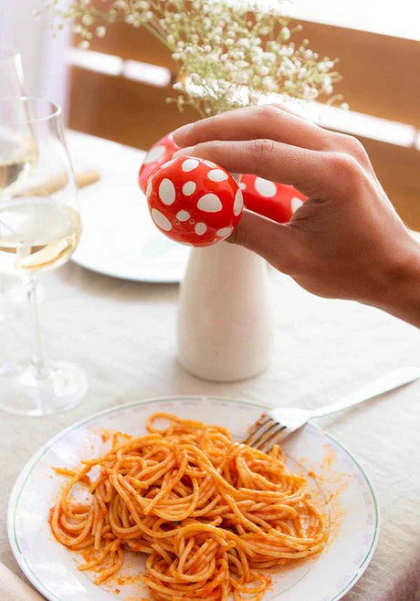 Hand holding red and white spotted Amanita mushroom salt shaker over plate of spaghetti on dining table