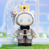 The Lucky EMMA - Secret Forest Wedding figurine by Lucky Emma features a crown and bow tie, a round face with colorful eyes, and stands on grass amidst a beautifully blurred background. Part of the Collectible Quality series, it's adorable for any collection.