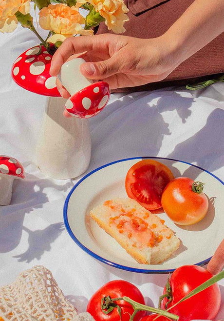 Hand holding red-and-white spotted mushroom salt shaker near plate with bread and tomatoes on white cloth