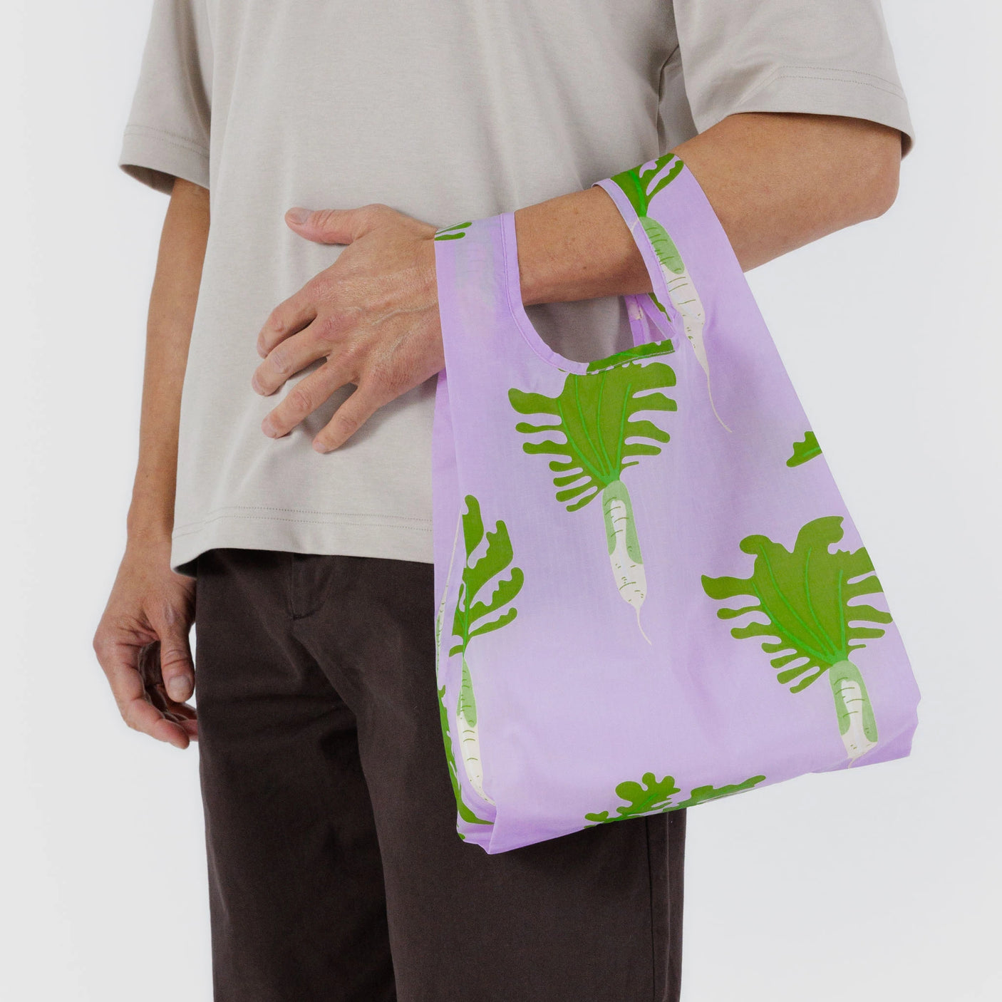 A person carries a Baggu Baby Baggu Reusable Bag in Daikon Pink, featuring green leaf and white radish designs, while wearing a light gray shirt and dark pants.