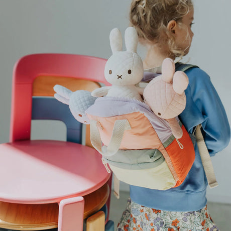 A child with a vibrant, handcrafted backpack filled with Miffy Quilted Plush Gift Box toys stands beside a pink and wooden chair.