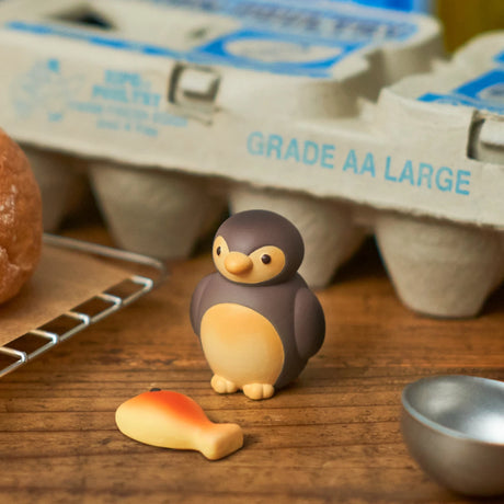 A small penguin figurine from the Animal Bakery Blind Box - Vol 2 by Dreams Inc and a fish-shaped object sit on a wooden surface, with an egg carton and other kitchen items in the background.