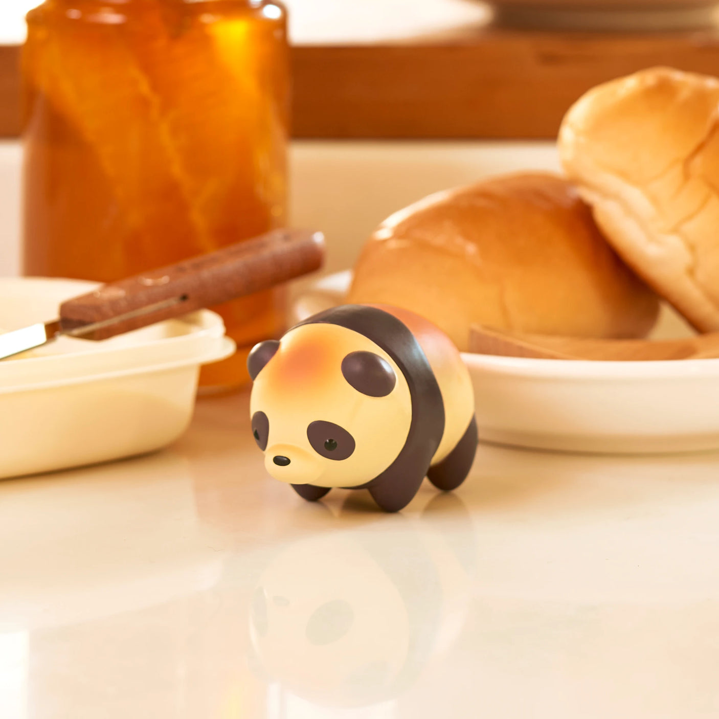 A small panda figurine from the Animal Bakery Mini Figure Blind Box by Dreams Inc sits on a kitchen counter next to bread and a jar.