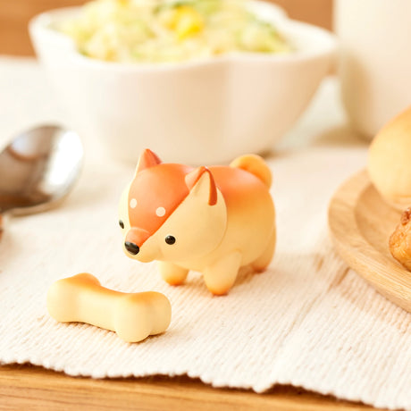 A charming dog figurine from Dreams Inc's Animal Bakery Mini Figure Blind Box series showcases its toy-like form with orange and cream colors, sitting beside a bone on a white tablecloth. Adding an extra touch of whimsy to this scene are a spoon, some bread, and a bowl of salad. Ideal for any collector or Blind Box fan.