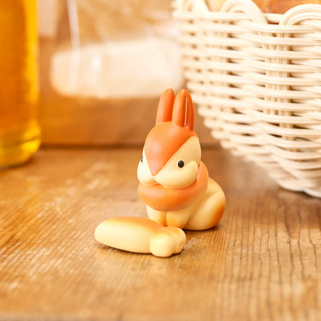 From the Animal Bakery Mini Figure Blind Box by Dreams Inc, a small, cartoon-style rabbit figurine decorates a wooden surface beside a loaf of bread in a wicker basket. It's perfect for adding charm to any Collectible Series display.