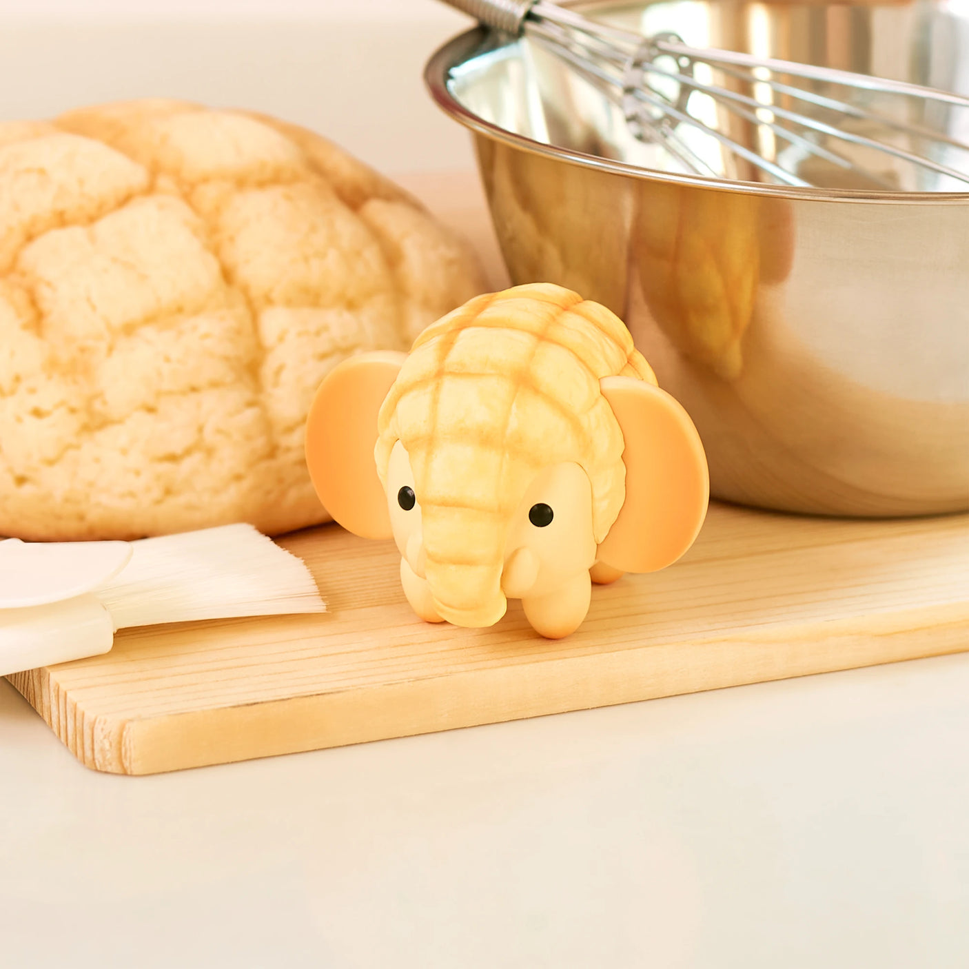 A charming Animal Bakery Mini Figure from the exclusive Dreams Inc collection, resembling a pastry, is displayed on a wooden board beside a loaf of similar bread, near a mixing bowl and whisk.