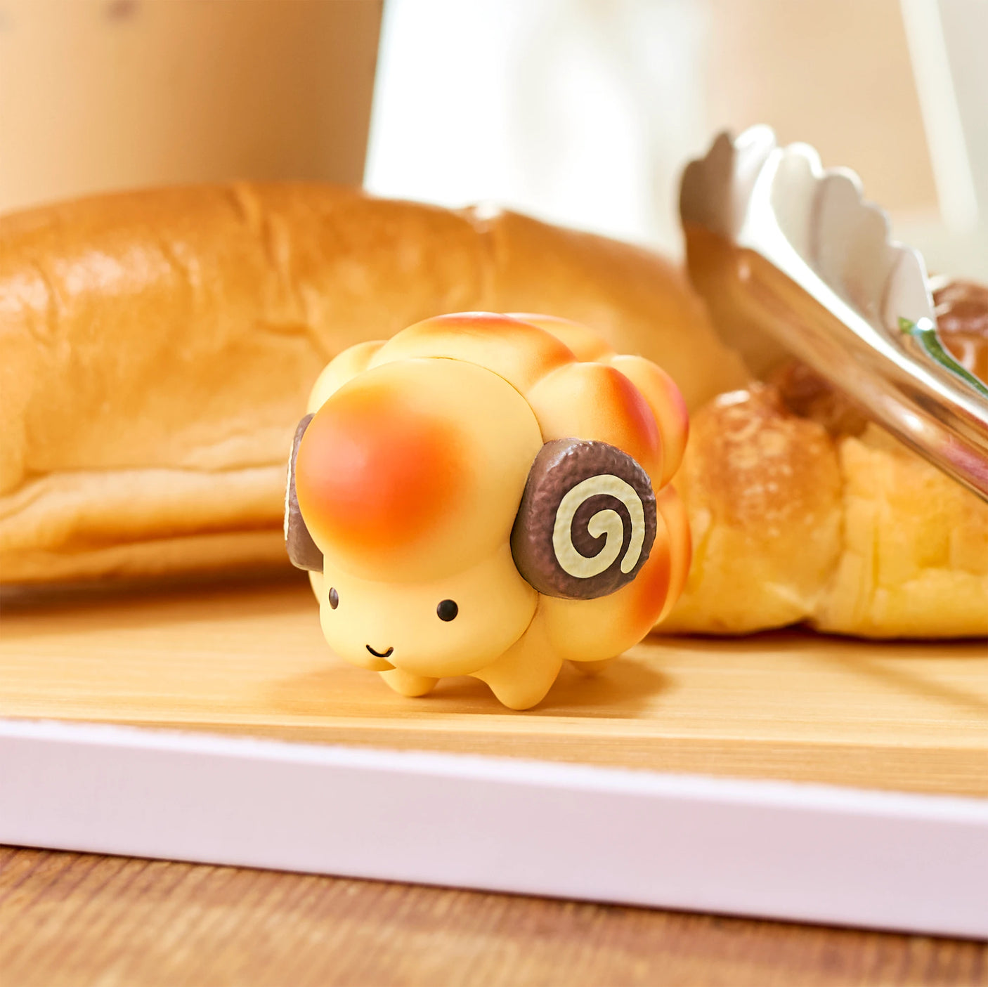 A delightful toy sheep featuring swirl-patterned horns is showcased on a wooden board, surrounded by fresh bread and pastries. This charming piece is part of Dreams Inc's Animal Bakery Mini Figure Blind Box series. Unwrap the mystery inside each box to bring a delightful surprise to your collection.