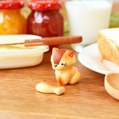 A tiny figurine from the Animal Bakery Mini Figure Blind Box collection by Dreams Inc is displayed on a wooden table beside a fish-shaped object. It is encircled by jars of jam, a butter dish, a glass of milk, and a slice of bread.