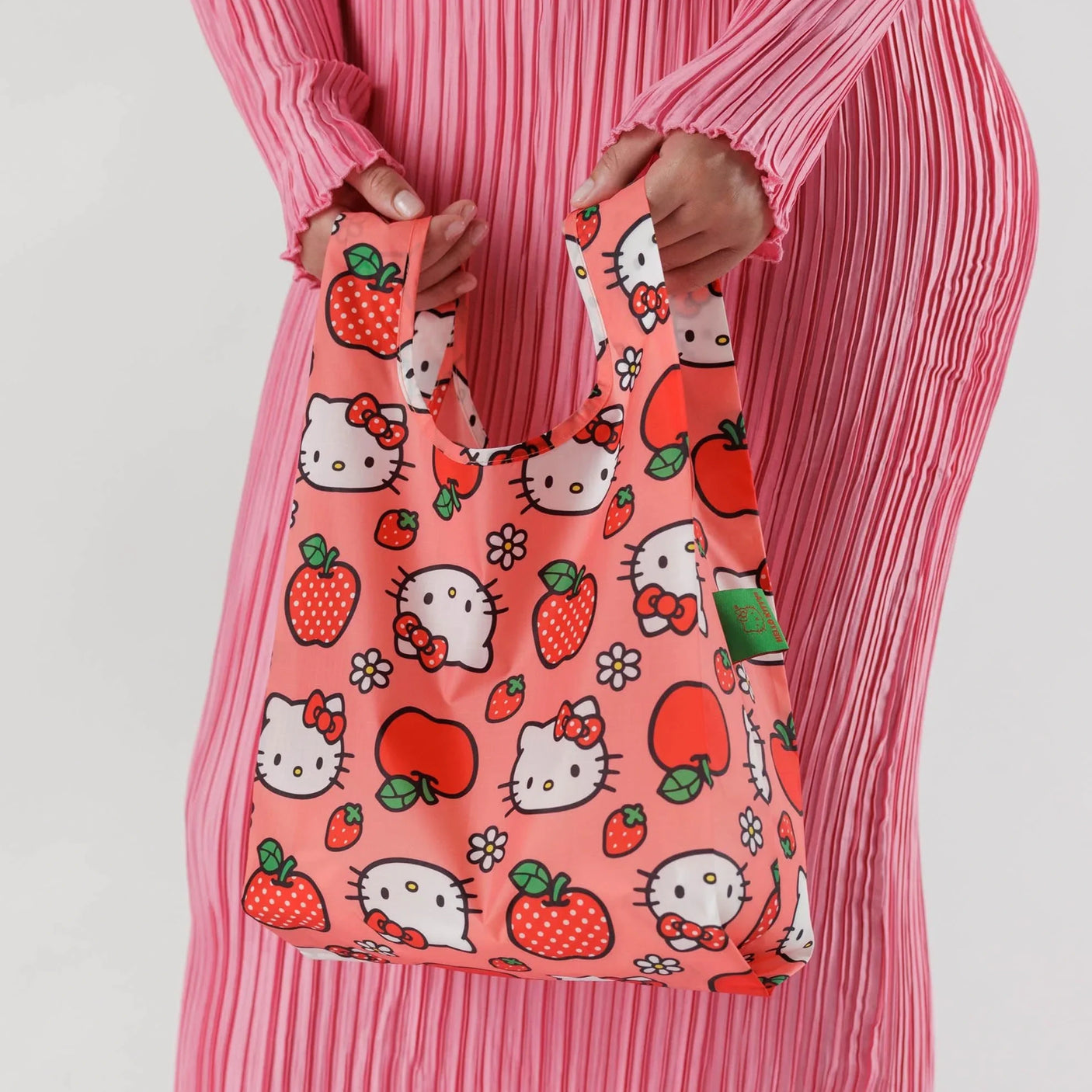 A person in a pink dress holds a limited edition Baggu Baby Baggu Bag - Hello Kitty Apple, featuring cartoon cat faces, strawberries, and apples.