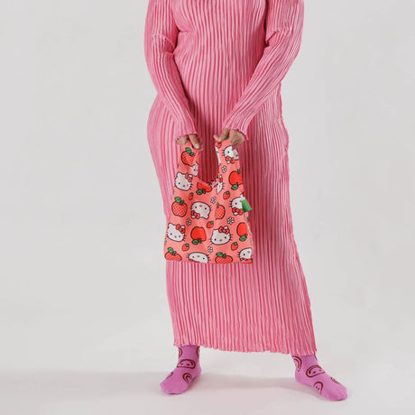 A person in a pink dress holds a limited edition Baby Baggu Bag adorned with Hello Kitty Apple cartoons. They are wearing matching pink socks against a white background.