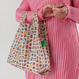 A person in a pink dress holds the Baby Baggu Bag - Hello Kitty Icons by Baggu, a limited edition tote featuring vibrant cartoon fruit, flowers, and Hello Kitty designs.