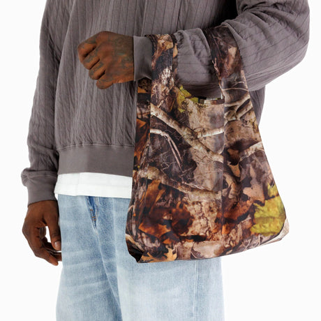 A person holds a tote in the "Photo Forest" design, similar to a Baby Baggu Reusable Bag from Baggu, alongside a gray quilted jacket and light blue jeans. This bag combines eco-friendly recycled nylon with an appealing aesthetic, ideal for those who value both style and sustainability.