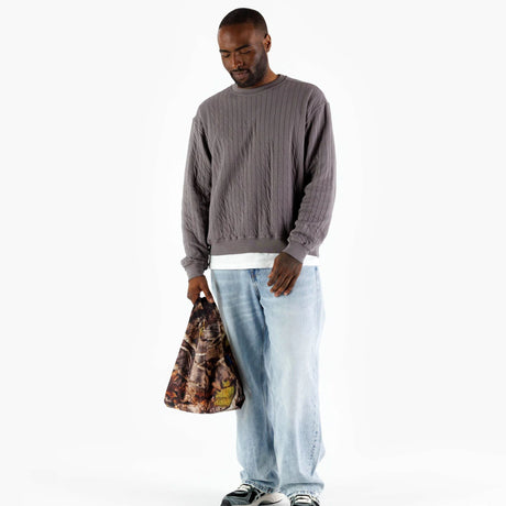 A person wearing a gray sweater and light denim jeans stands holding a Baggu Baby Baggu reusable bag in the Photo Forest design, made from recycled nylon.