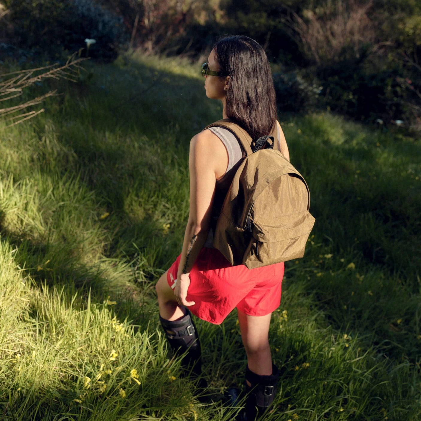 A person wearing sunglasses, a sleeveless top, and red shorts stands on a grassy path with sunlight filtering through the trees. They carry a Baggu Medium Nylon Backpack in Digital Denim, blending style and sustainability into their adventurous ensemble.