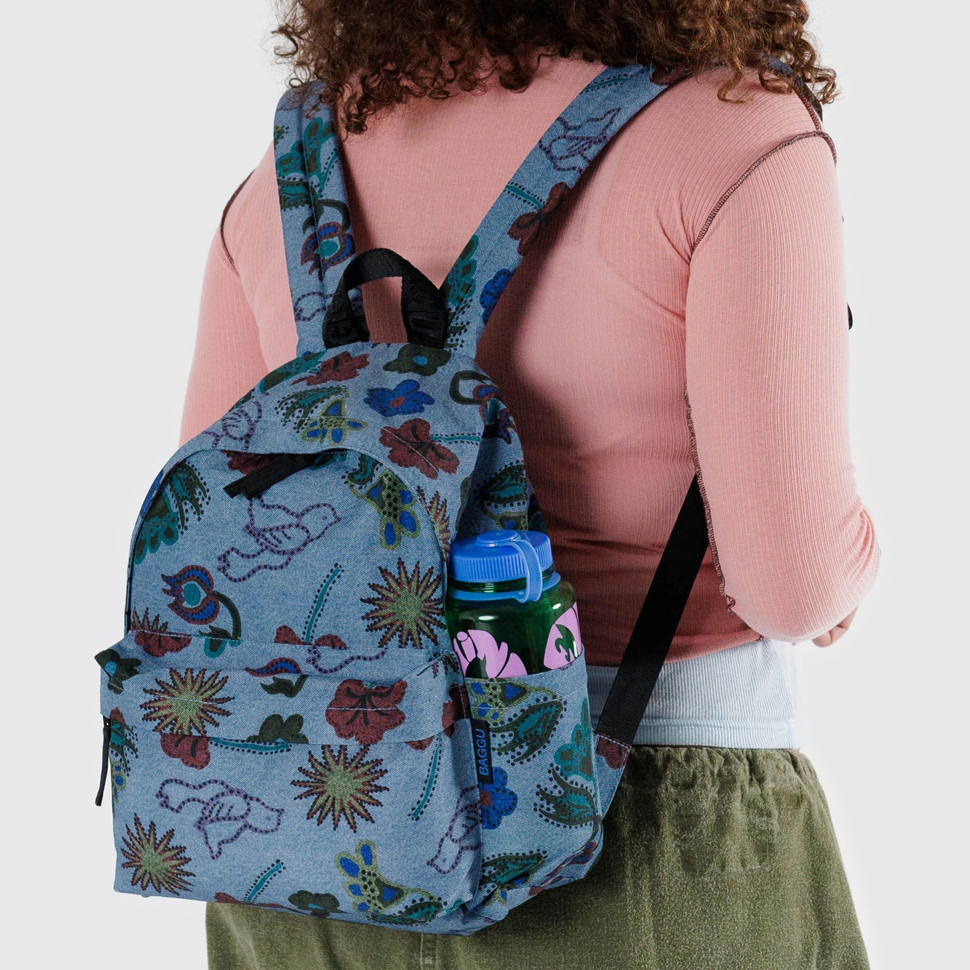 A person strolls with a Baggu Medium Nylon Backpack in Digital Denim, beautifully complemented by floral patterns. The pink water bottle fits snugly into the side pocket, showcasing sustainable style with its construction from recycled nylon.