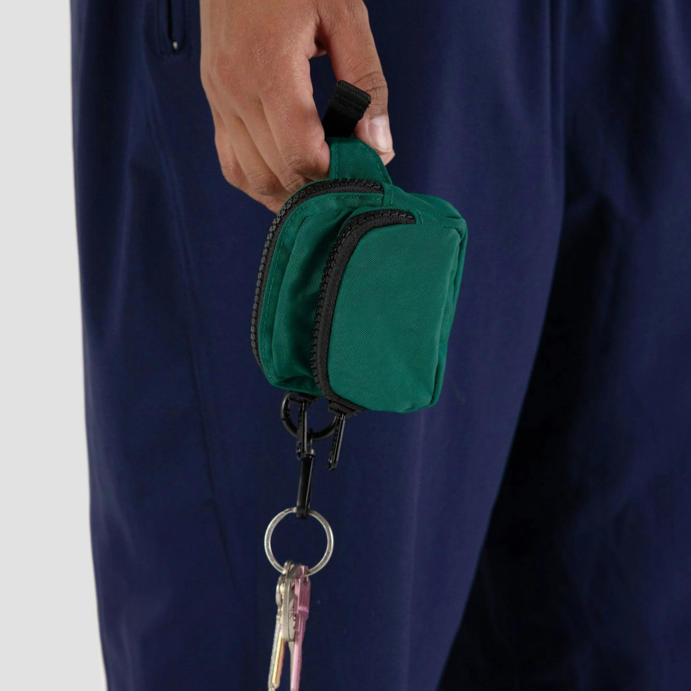 A hand holds the Baggu Fanny Pack Charm - Cypress, crafted from recycled nylon and attached to a keychain with keys. Its vibrant green contrasts beautifully against navy blue pants, adding a stylish touch for those on the go.