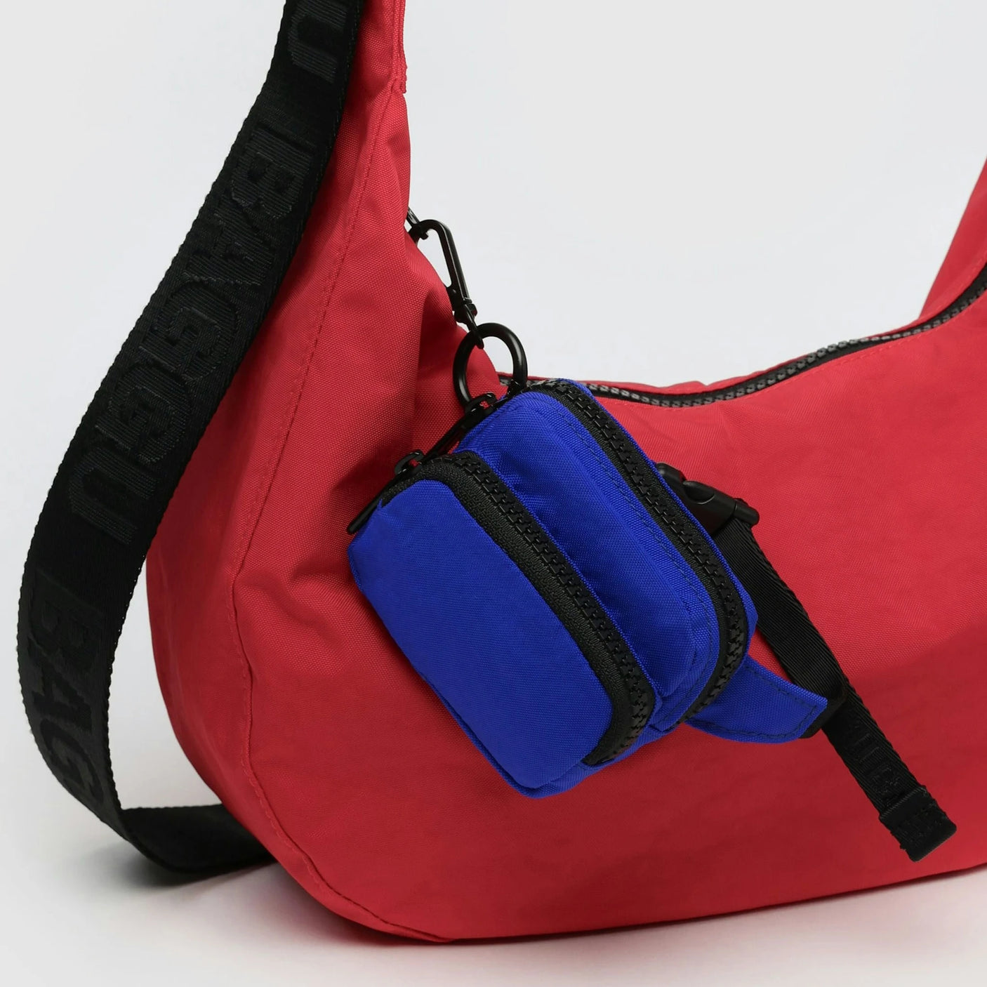 The Baggu Fanny Pack Charm is a red shoulder bag made from recycled heavyweight nylon, featuring a black strap and an attached small blue pouch. Its playful design includes an additional fanny pack charm for flair and functionality.