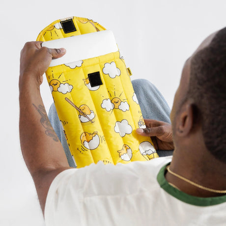 A person holds a vibrant yellow item featuring playful cloud and egg designs reminiscent of Gudetama, set against a crisp white background from the Gudetama x Baggu Puffy Laptop Sleeve collection.