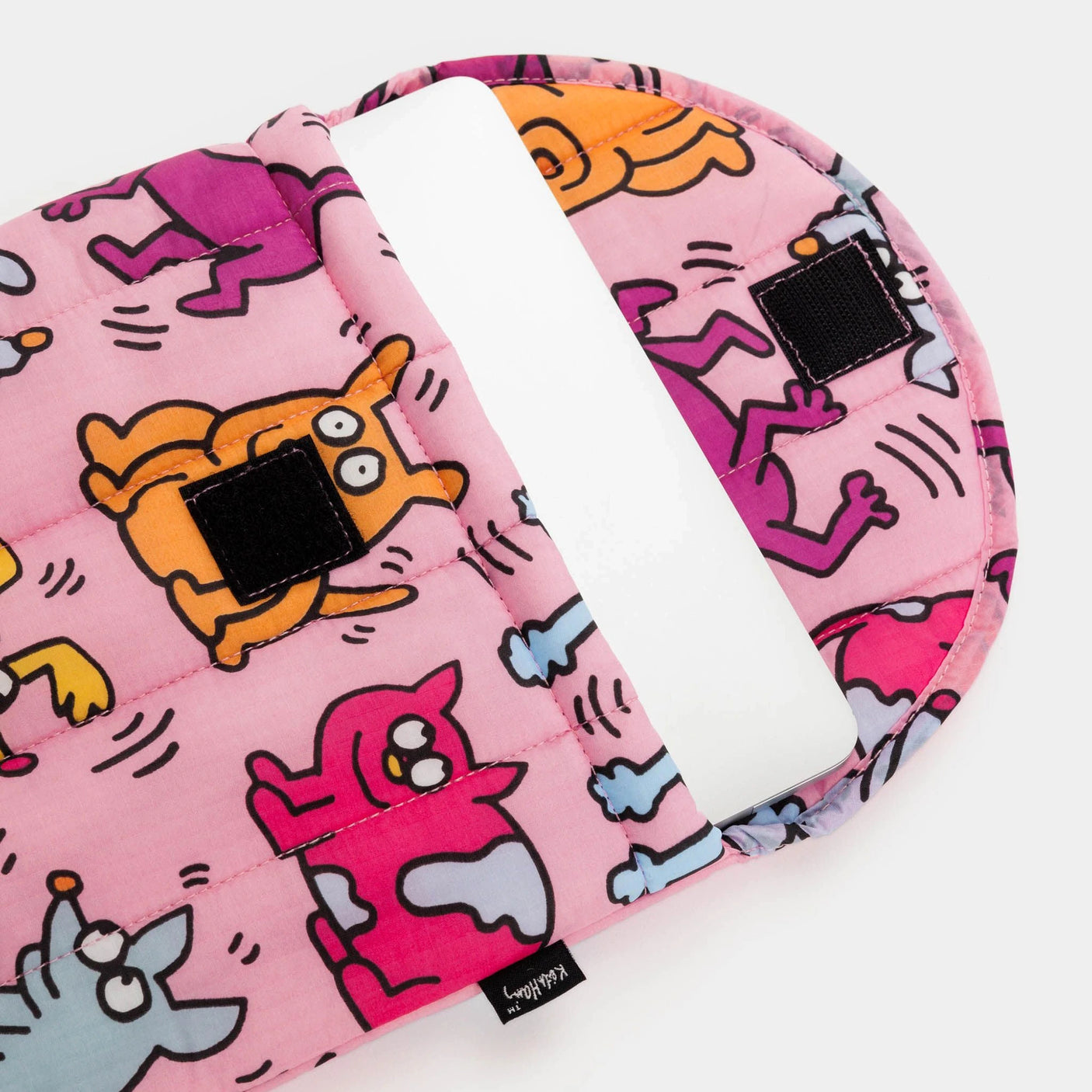 A Keith Haring x Baggu Puffy Laptop Sleeve in pink showcases playful Pets designs and includes a secure strap, partially open to reveal a white tablet inside. This eco-friendly case offers enhanced protection with its puffy design, while incorporating recycled materials.