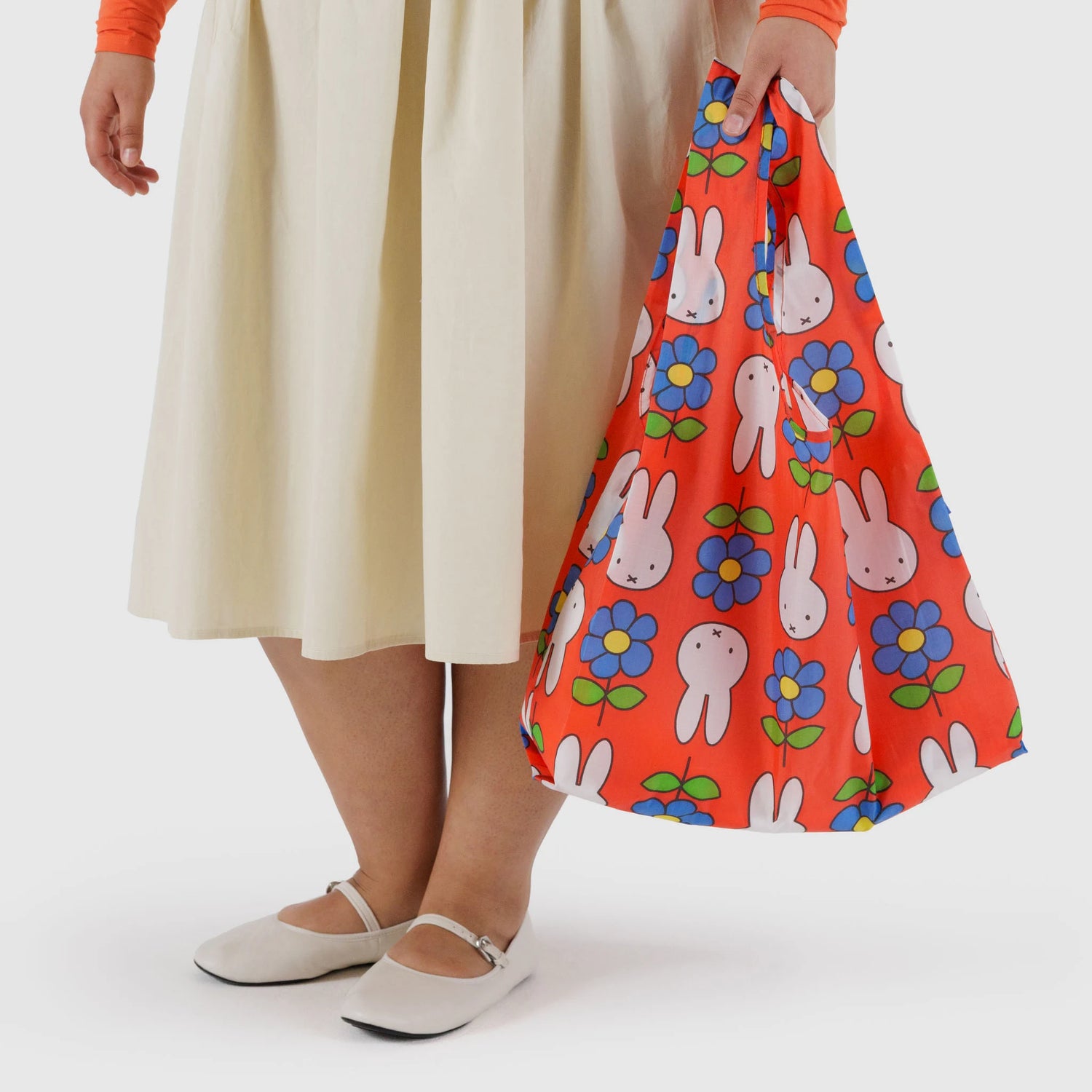 A person in a cream skirt, white shoes, and orange top holds the Baggu Standard Baggu x Miffy Flower— a red reusable bag featuring bunny and blue flower patterns.