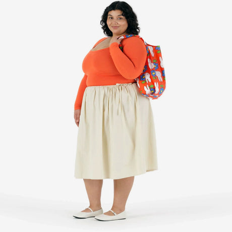A woman in an orange long-sleeve top, beige midi skirt, and white shoes stands holding the Baggu Standard Baggu x Miffy Flower reusable bag over her shoulder, set against a plain white background.