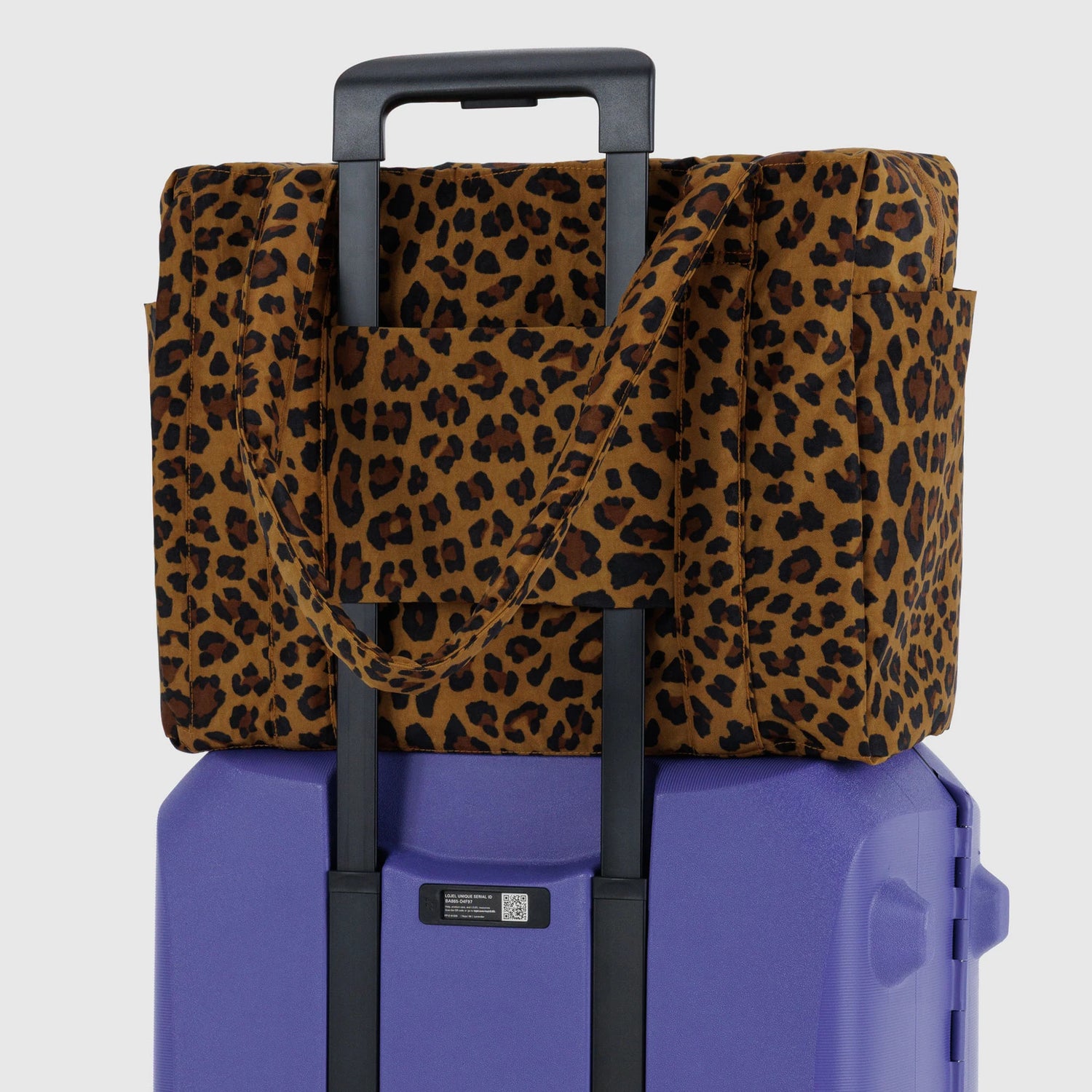 The Baggu Small Cloud Carry-on in Leopard by Baggu sits securely atop a purple rolling suitcase, attached via the handle for a stylish and lightweight travel setup.
