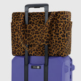 The Baggu Small Cloud Carry-on in Leopard by Baggu sits securely atop a purple rolling suitcase, attached via the handle for a stylish and lightweight travel setup.