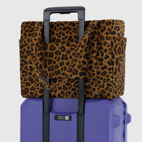 The Baggu Small Cloud Carry-on in Leopard by Baggu sits securely atop a purple rolling suitcase, attached via the handle for a stylish and lightweight travel setup.