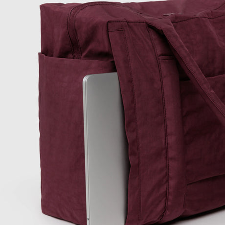 A silver laptop peeks from the side pocket of the Baggu Small Cloud Carry-on in Mahogany by Baggu, set against a plain background.