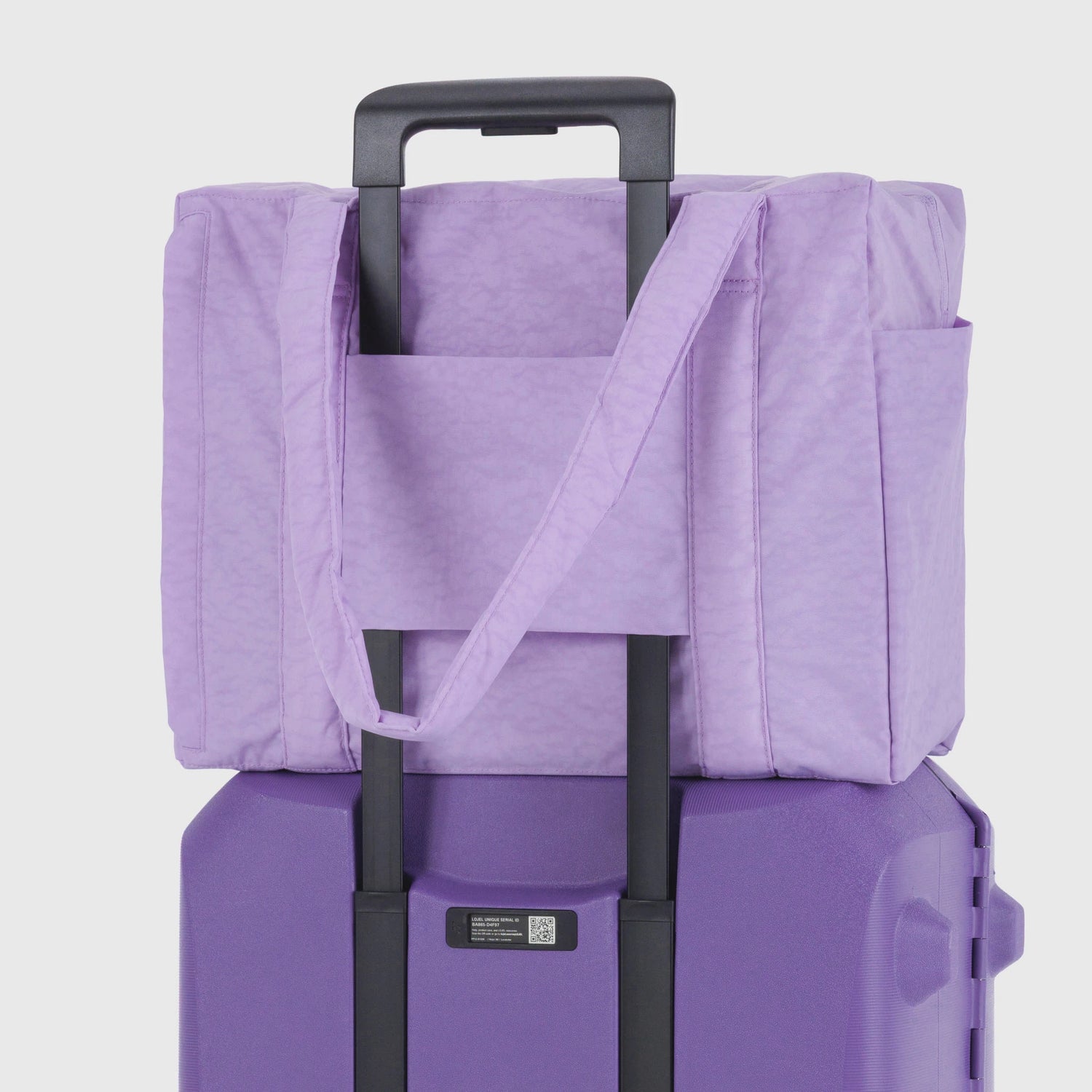 The Baggu Small Cloud Carry-on in Wisteria is shown securely placed over the handle of a purple rolling suitcase by Baggu, set against a plain backdrop. Perfect as a travel carry-on.