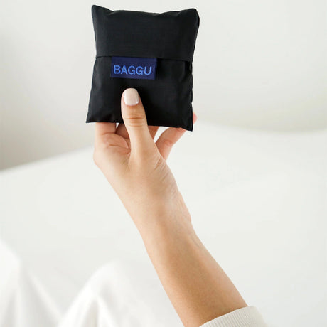 A hand holds a small, folded Baby Baggu Reusable Bag - Black from Baggu, crafted from recycled nylon, against a light background.