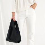 Dressed in a sleek white outfit, a person elegantly holds a Baby Baggu Reusable Bag in black from Baggu, highlighting their chic yet eco-conscious style.