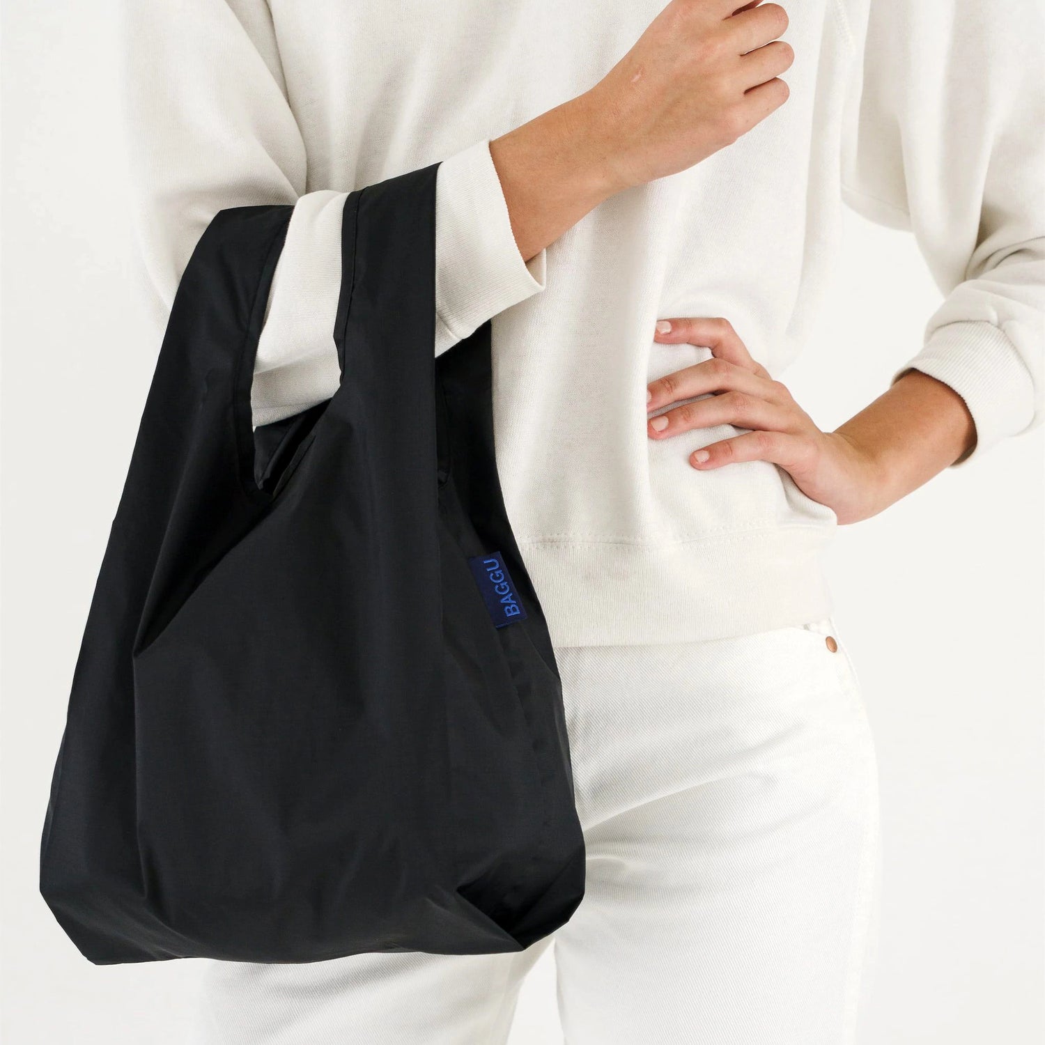 A person dressed in white carries a stylish black Baby Baggu Reusable Bag from the brand Baggu, made from recycled nylon, on their arm.