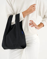 A person dressed in white carries a stylish black Baby Baggu Reusable Bag from the brand Baggu, made from recycled nylon, on their arm.