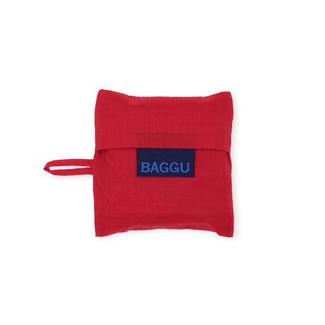 The Candy Apple Baby Baggu Reusable Bag by Baggu is a stylish and sustainable red fabric pouch, made from 100% recycled nylon. It features a handle, folds neatly, and is adorned with a blue label that reads "BAGGU.