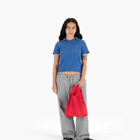 A person in a blue shirt and grey pants holds a Baggu Baby Baggu Reusable Bag in Candy Apple red against a neutral background.