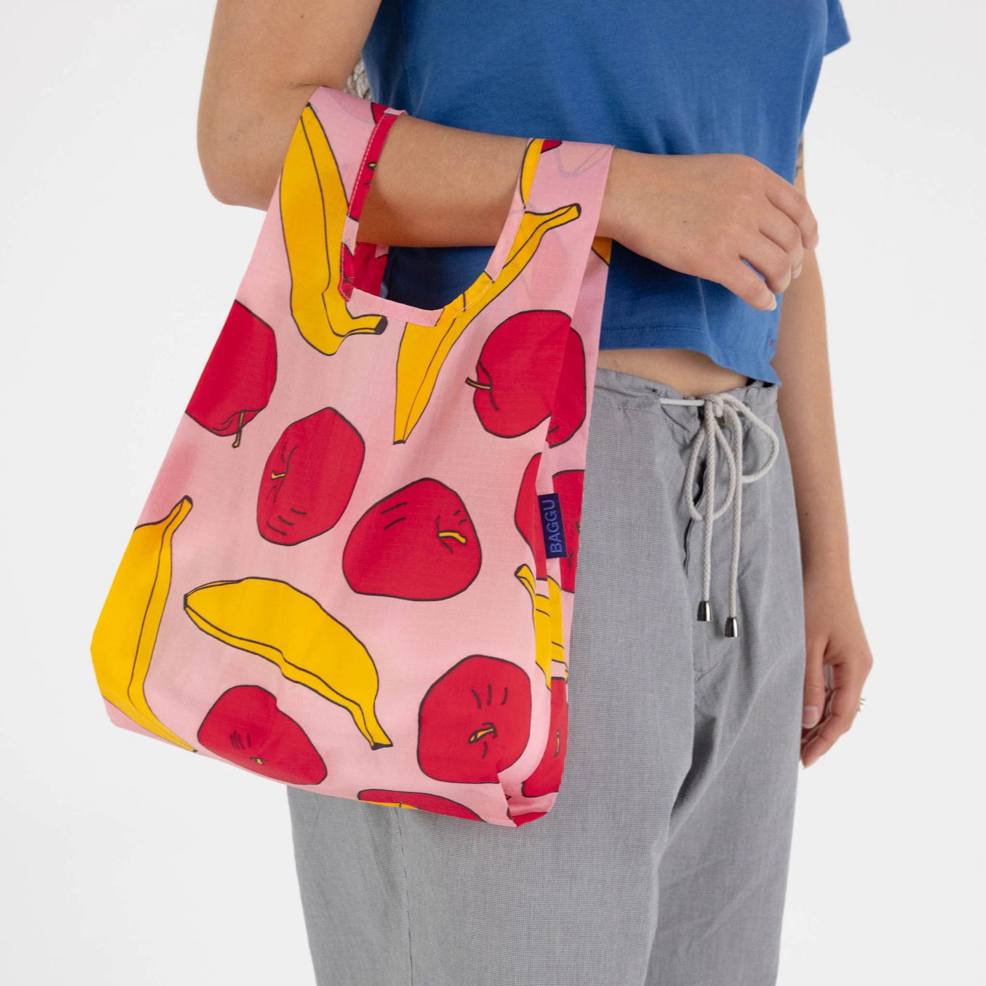 A person carries a Baby Baggu Reusable Bag in pink, featuring apples and bananas from the brand Baggu, filled with red apples and yellow bananas, while wearing a blue top and gray pants.