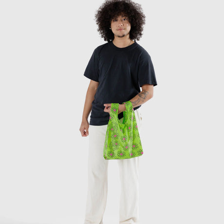 A person with curly hair, dressed in a black shirt and white pants, holds a Baby Baggu reusable bag from Baggu featuring the Keith Haring Flower design.