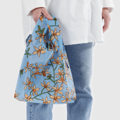 A person holds a Baby Baggu Reusable Bag - Orchid, featuring a delicate orchid print and crafted from 100% recycled nylon by Baggu.