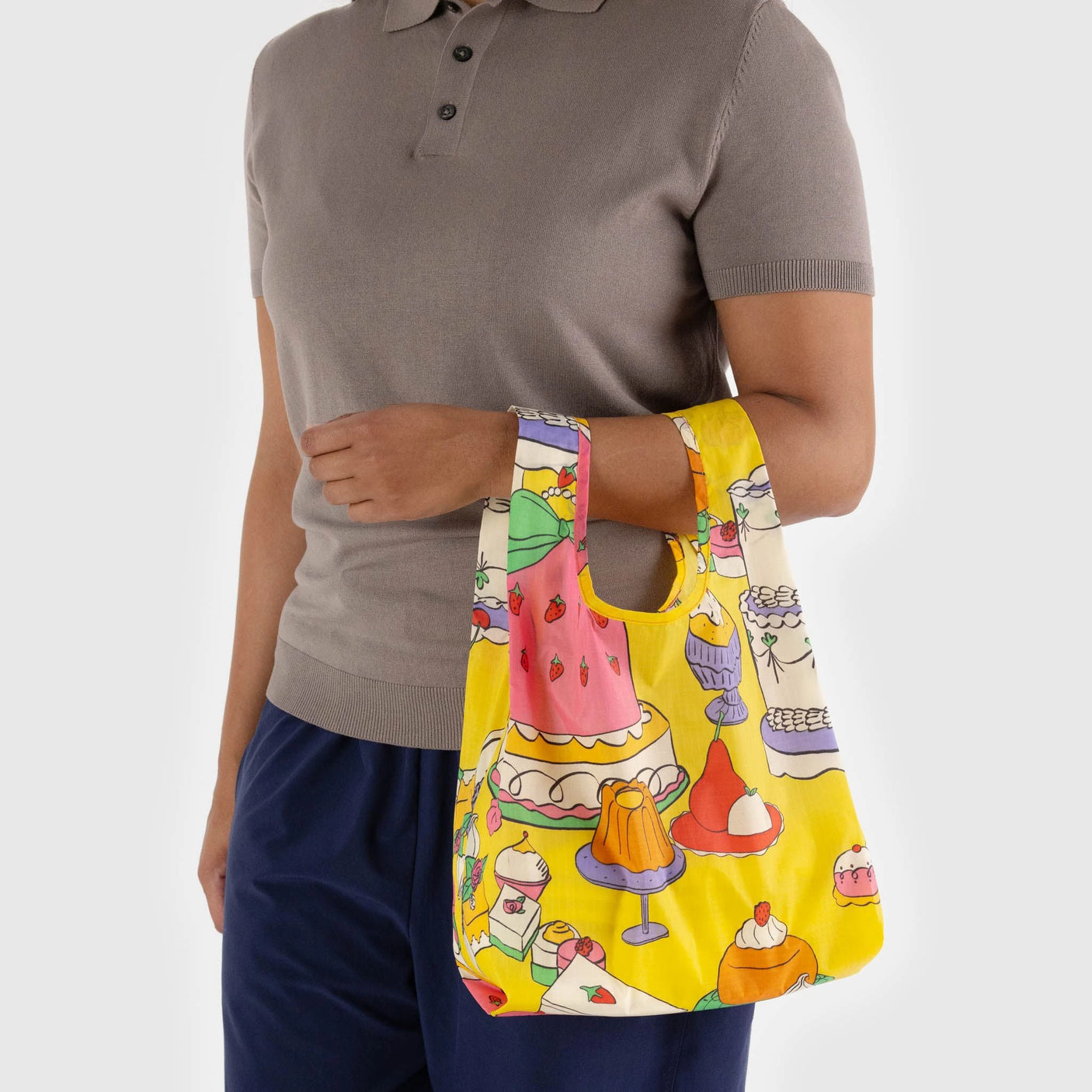 Wearing a gray shirt and navy pants, a person displays a colorful Baby Baggu Reusable Bag - Patisserie by Baggu.