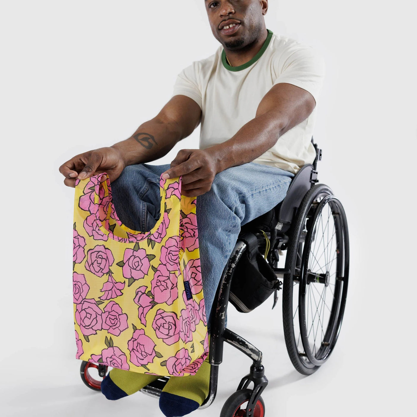 A man in a wheelchair holds a Baby Baggu Reusable Bag - Rose by Baggu, featuring a pink floral pattern and made from recycled nylon.