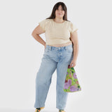 A person in a yellow top and jeans is holding the Baby Baggu Reusable Bag - Dahlia by Baggu against a plain background.