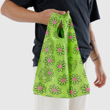 A person holds a Baby Baggu Keith Haring - Flower bag from Baggu, featuring pink flowers and black abstract patterns, while wearing a dark shirt and light pants.