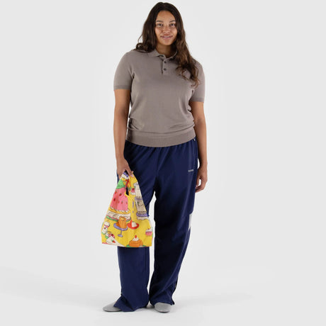 In front of a simple backdrop, a person stands dressed in a brown polo shirt and blue pants, carrying a bright yellow Baggu Baby Baggu Reusable Bag featuring lively patisserie designs.