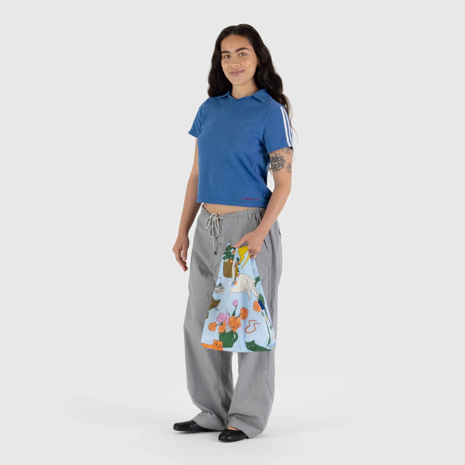 A person wearing a blue shirt and gray pants holds a Baby Baggu Reusable Bag - Table Cats from Baggu, featuring vibrant patterns, while standing against a plain background.